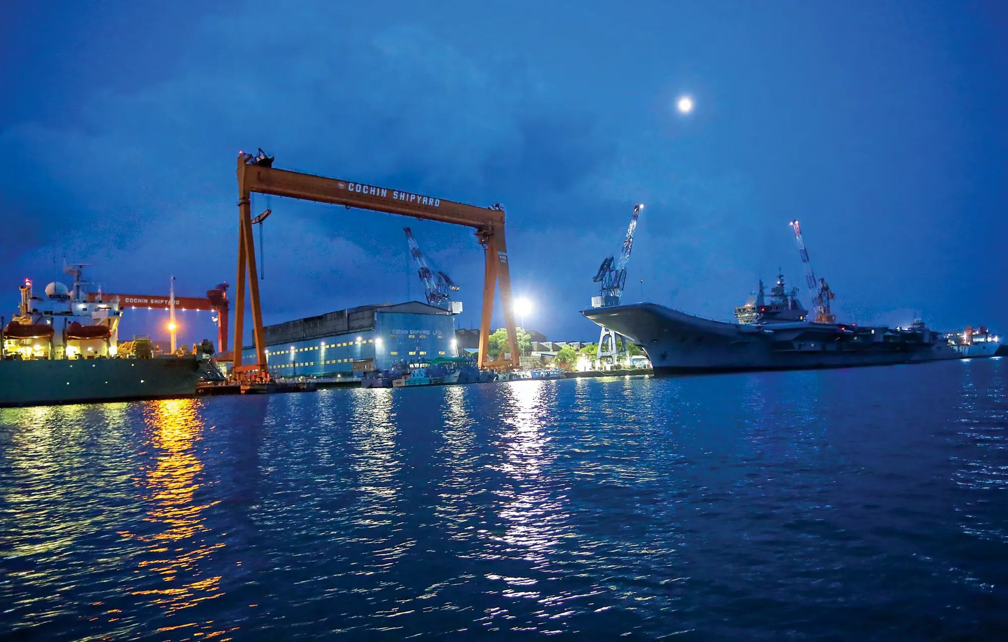 A warship alongside a shipyard gantry at dusk, the yard lit for night working.